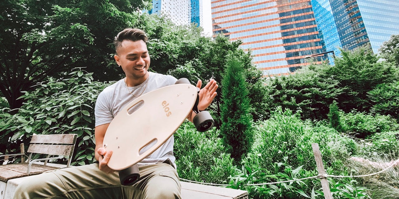 A Man holding maple Elos skateboards in the park of New York City, looking fulfilled after using skateboards for commuting