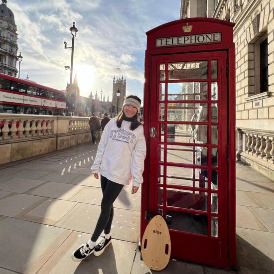 A girl smiling and standing beside an iconic London phone booth with her Elos skateboards nearby