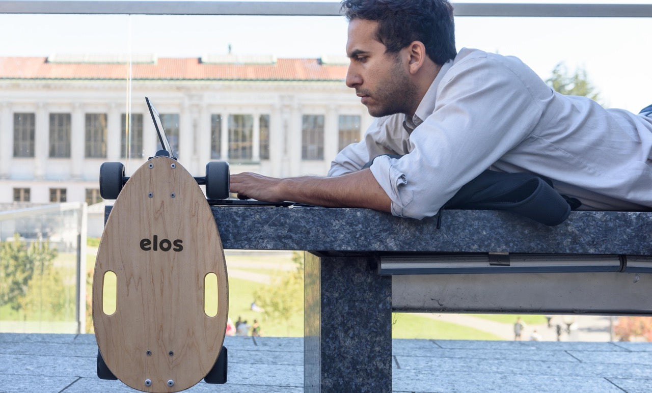 A man laying on the seat using his laptop while Elos is hanging next to him